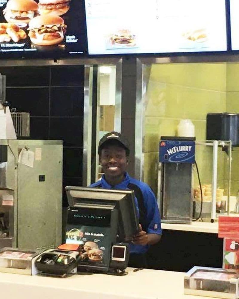 Holly working behind the counter at a McDonald's register, wearing a uniform cap and headset, smiling at the camera