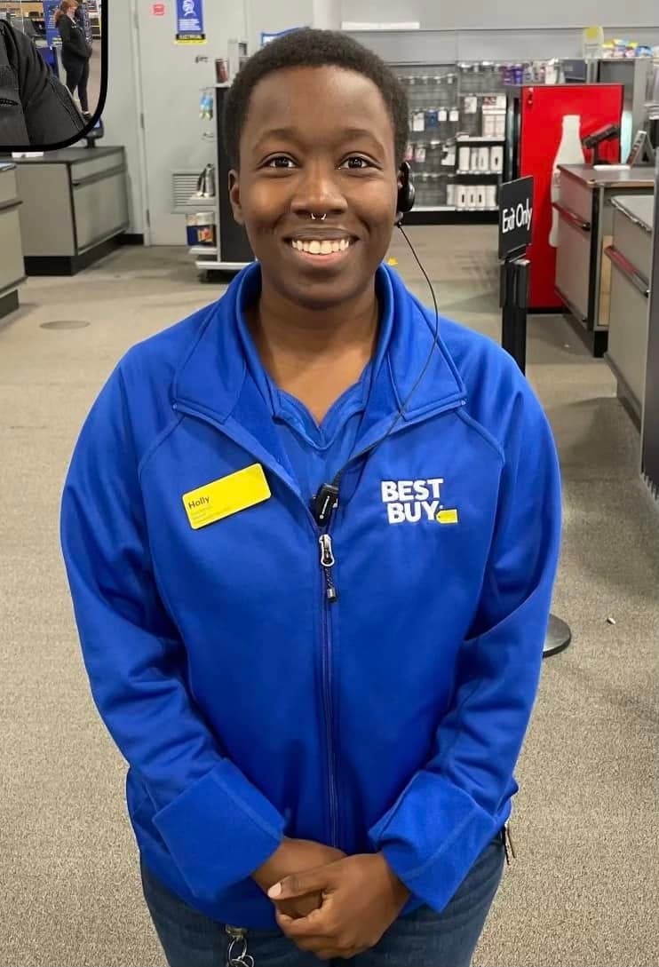 Holly smiling in a blue Best Buy jacket and headset, wearing a name tag, on the sales floor